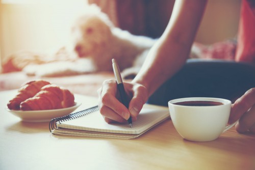female hands with pen writing on notebook with morning coffee and croissant